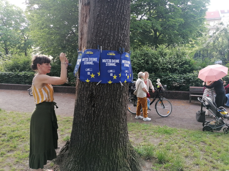 Frau hängt Plakate mit Europa-Fahne und "Nutze Deine Stimme" an Baumstamm
